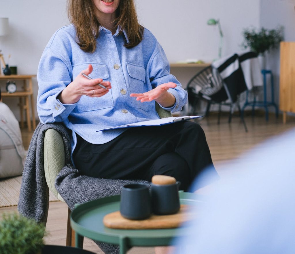 a woman with medium length brown hair in a blue shirt is speaking and gesturing with her hands with a notepad balanced on her knee