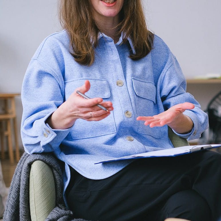 a woman with medium length brown hair in a blue shirt is speaking and gesturing with her hands with a notepad balanced on her knee