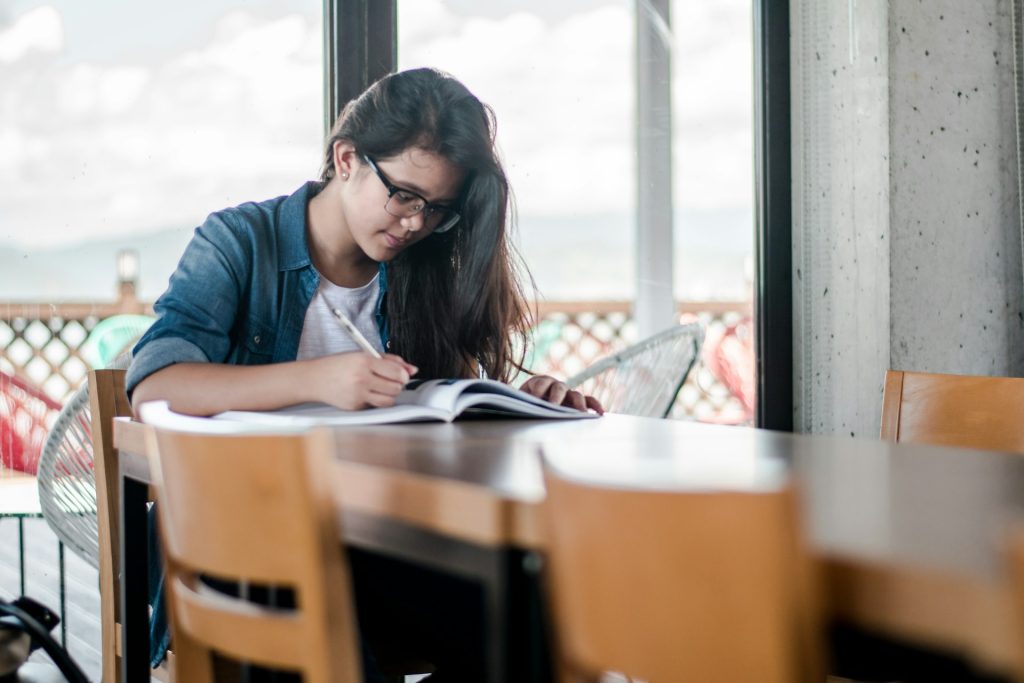 A young woman with long black hair and thich black rimmed glasses is sitting at a long table alone writing in a large exercise book. 