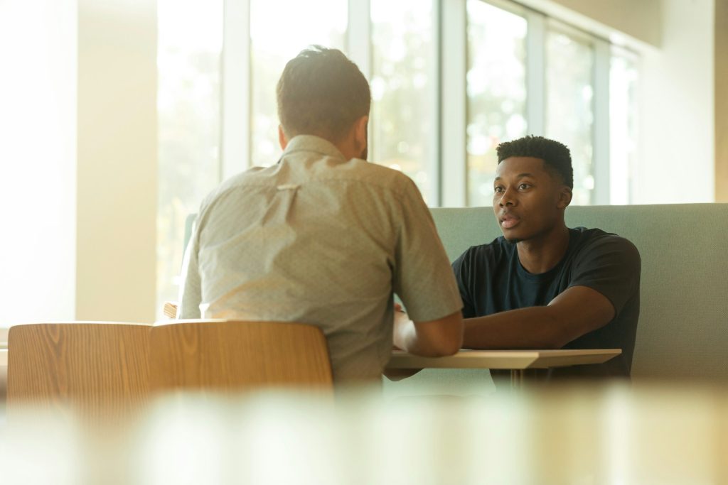 Two men talking across a table, a young black man is facing the camera.