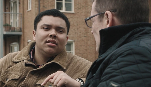 A young man speaking to an older man while they're both sat on a bench. The young man is facing the camera and has short black hair and is wearing a brown jacket. The older man has short brown hair, is wearing glasses and a back puffa jacket.