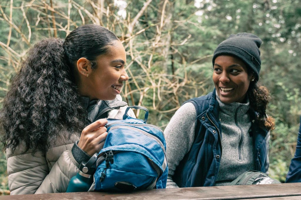 two women sat at a bench in the woods smiling at one another and talking