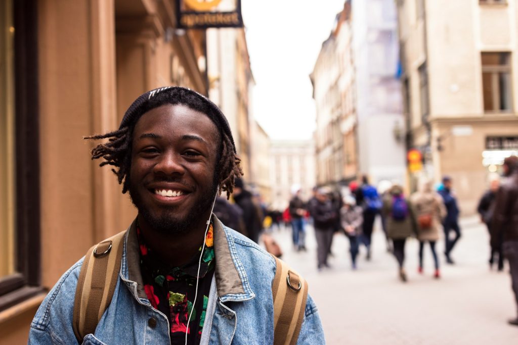 a young black man smiles at the camera on a busy street, he's wearing a black shirt with colourful shapes and a denim jacket